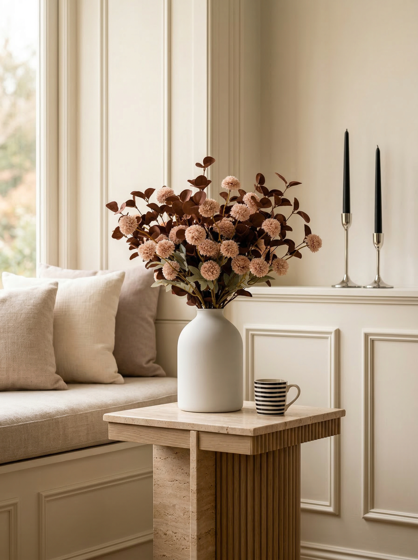 Decorative setup with a vase of flowers, candles, and a cup on a wooden table in a room with wainscoting.