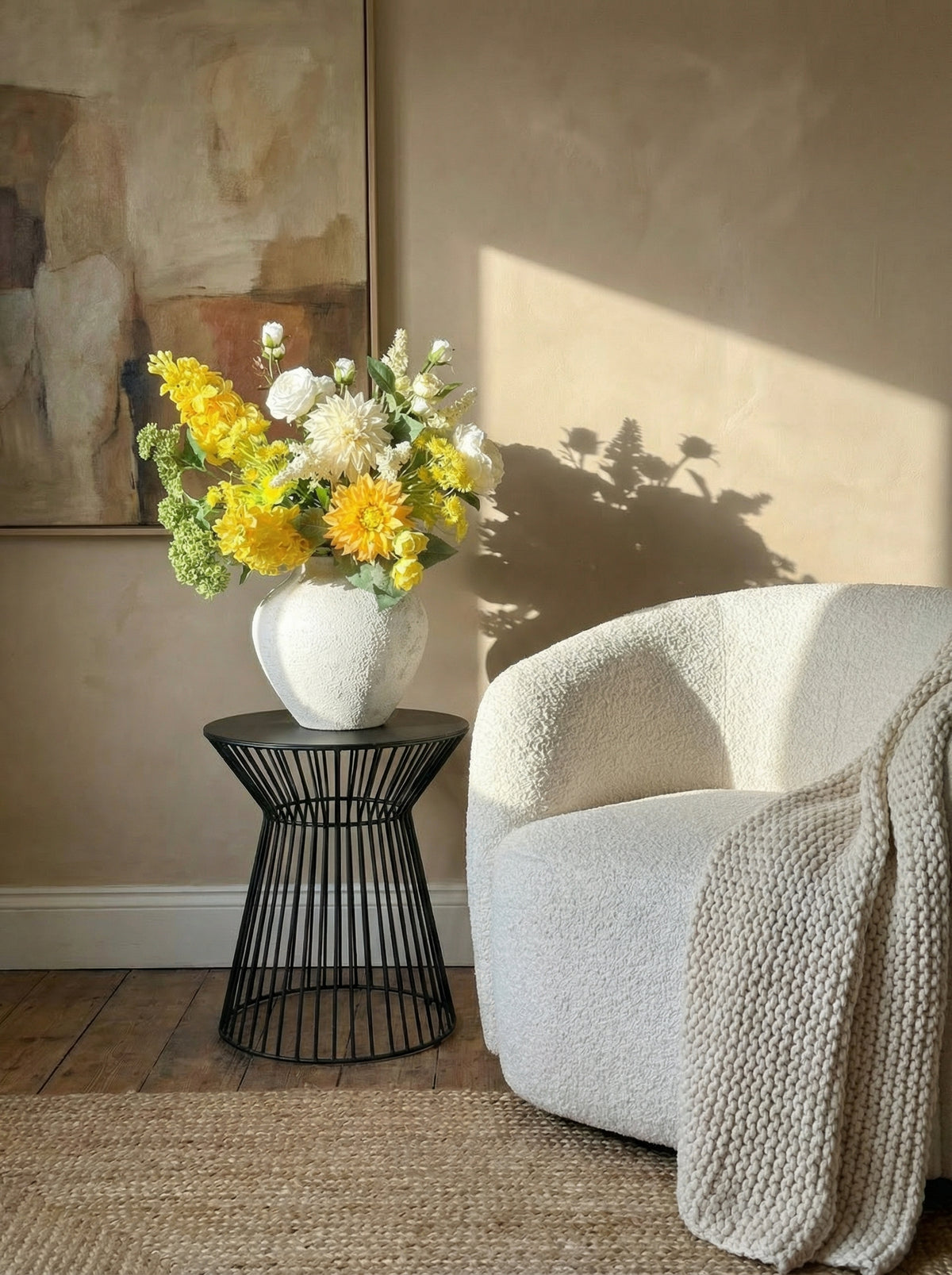 White vase with yellow and white flowers on a black side table next to a white armchair with a beige blanket.
