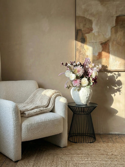 Beige armchair with a blanket and a vase of flowers on a small table in a room with a mirror.