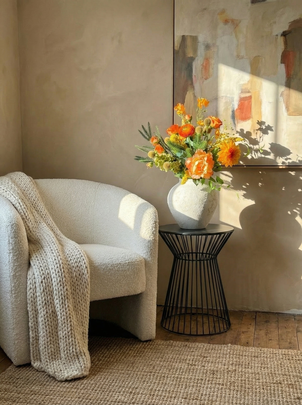 Beige armchair with a textured blanket, black side table with a vase of flowers, and abstract painting on a beige wall.