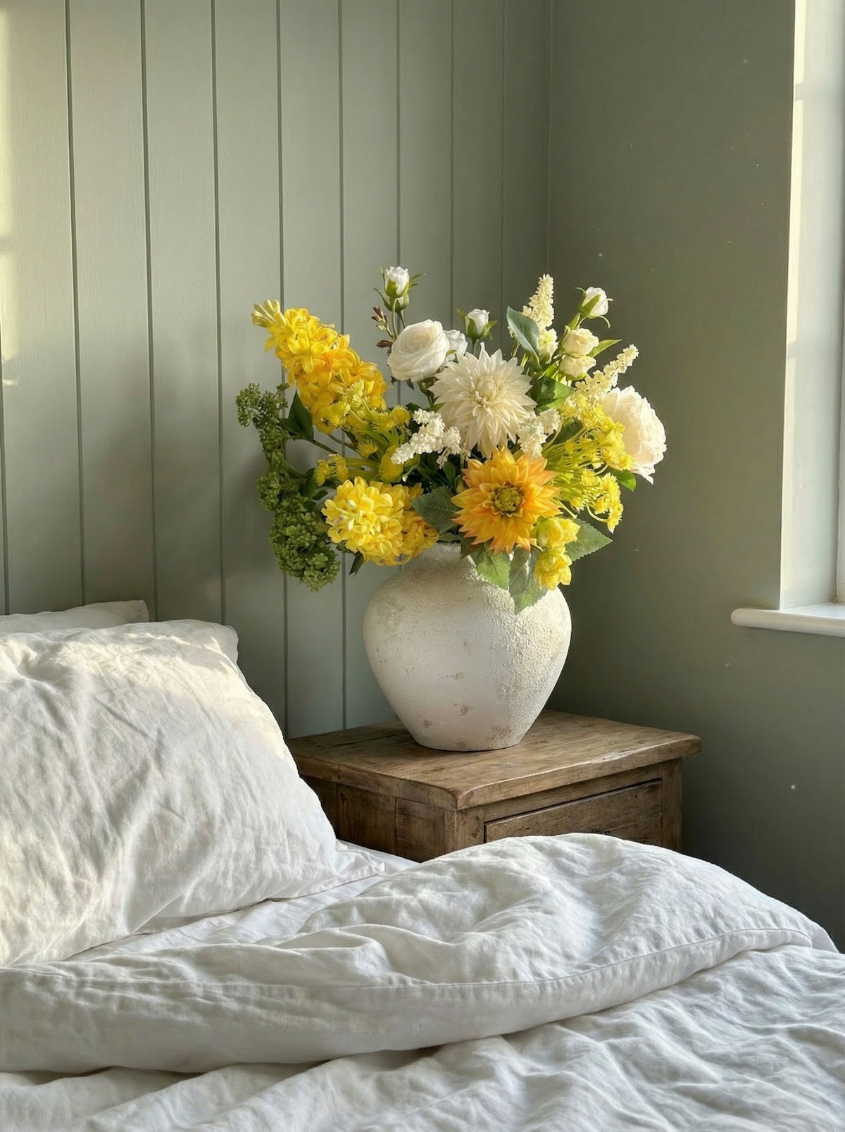 Bouquet of flowers in a vase on a wooden nightstand next to a bed with white bedding.
