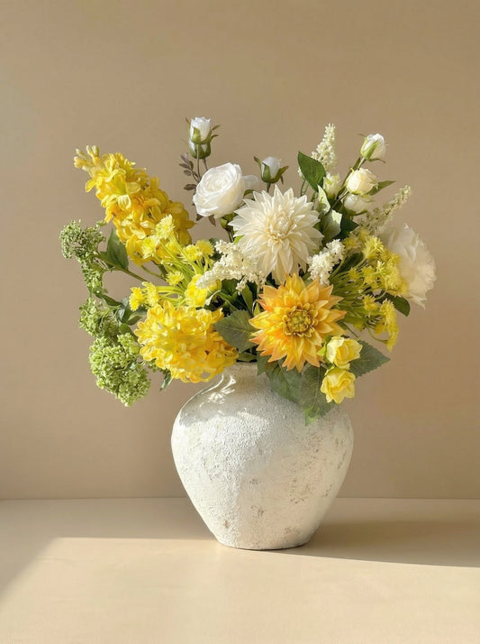Bouquet of yellow and white flowers in a textured white vase on a beige surface with a beige background