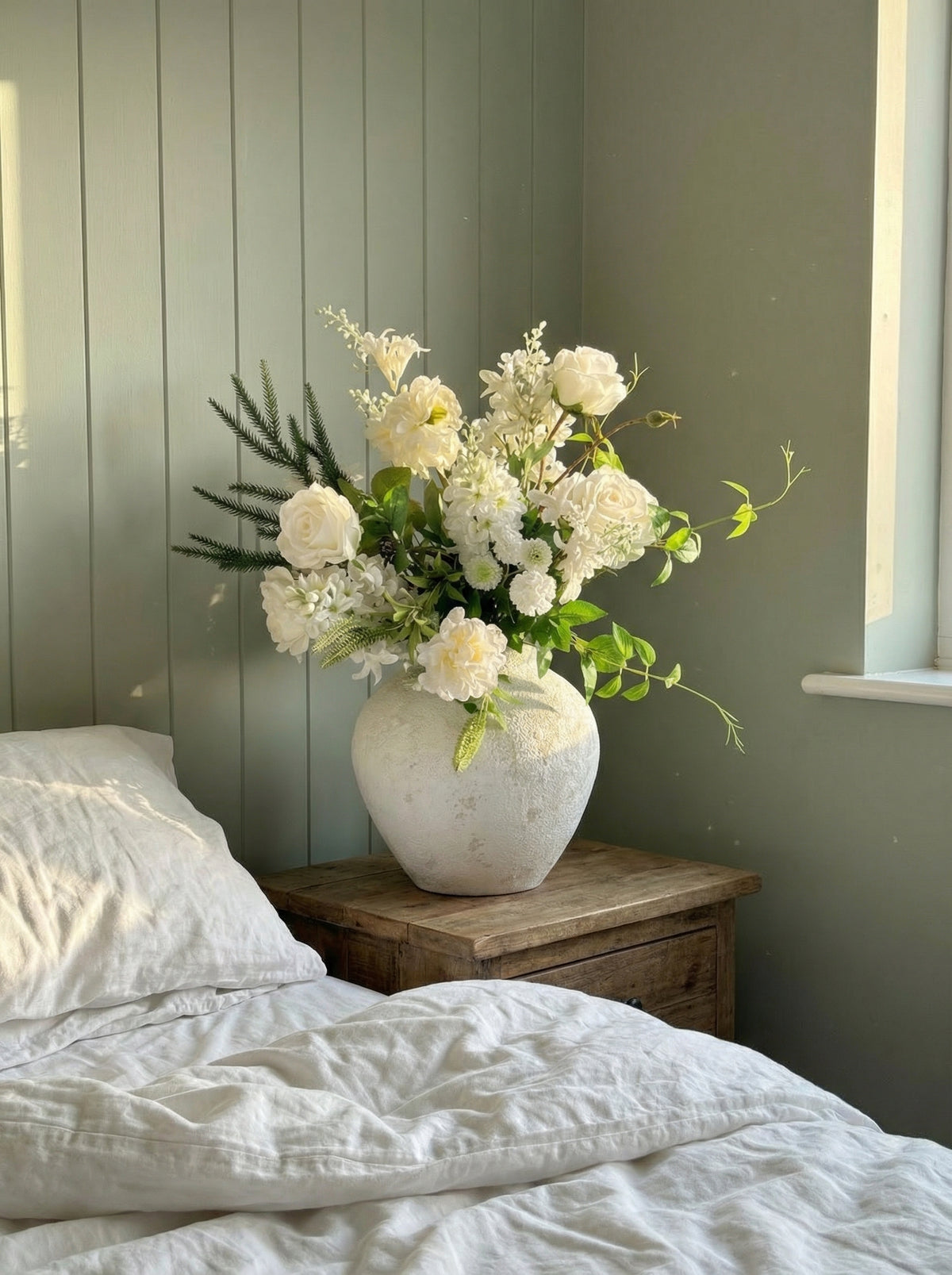 Floral arrangement in a textured vase on a wooden nightstand next to a bed with white bedding.