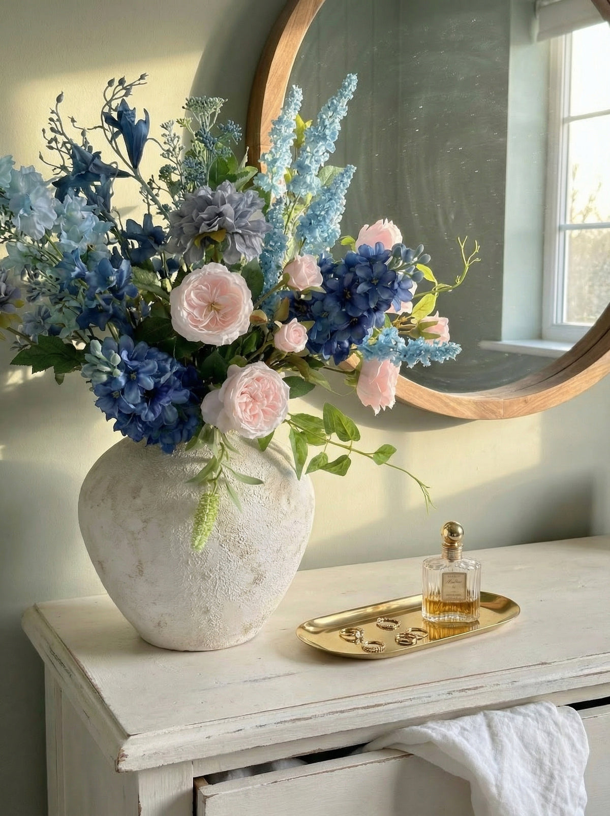 Floral arrangement in a textured vase on a wooden surface with a mirror and perfume bottle in the background.