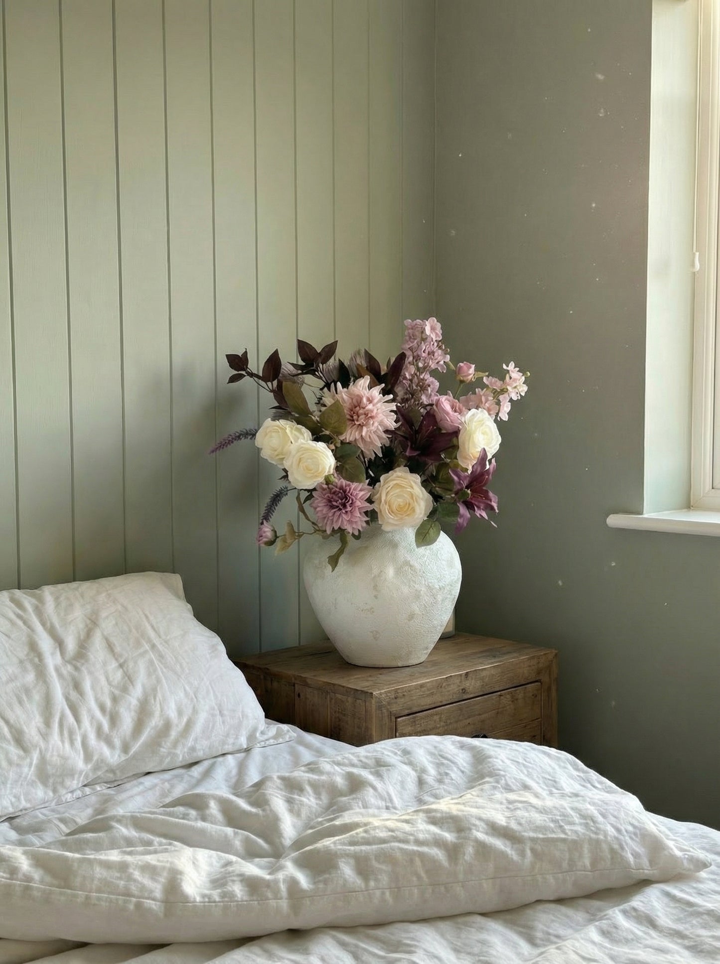 Floral arrangement in a white vase on a wooden nightstand next to a bed with white bedding.