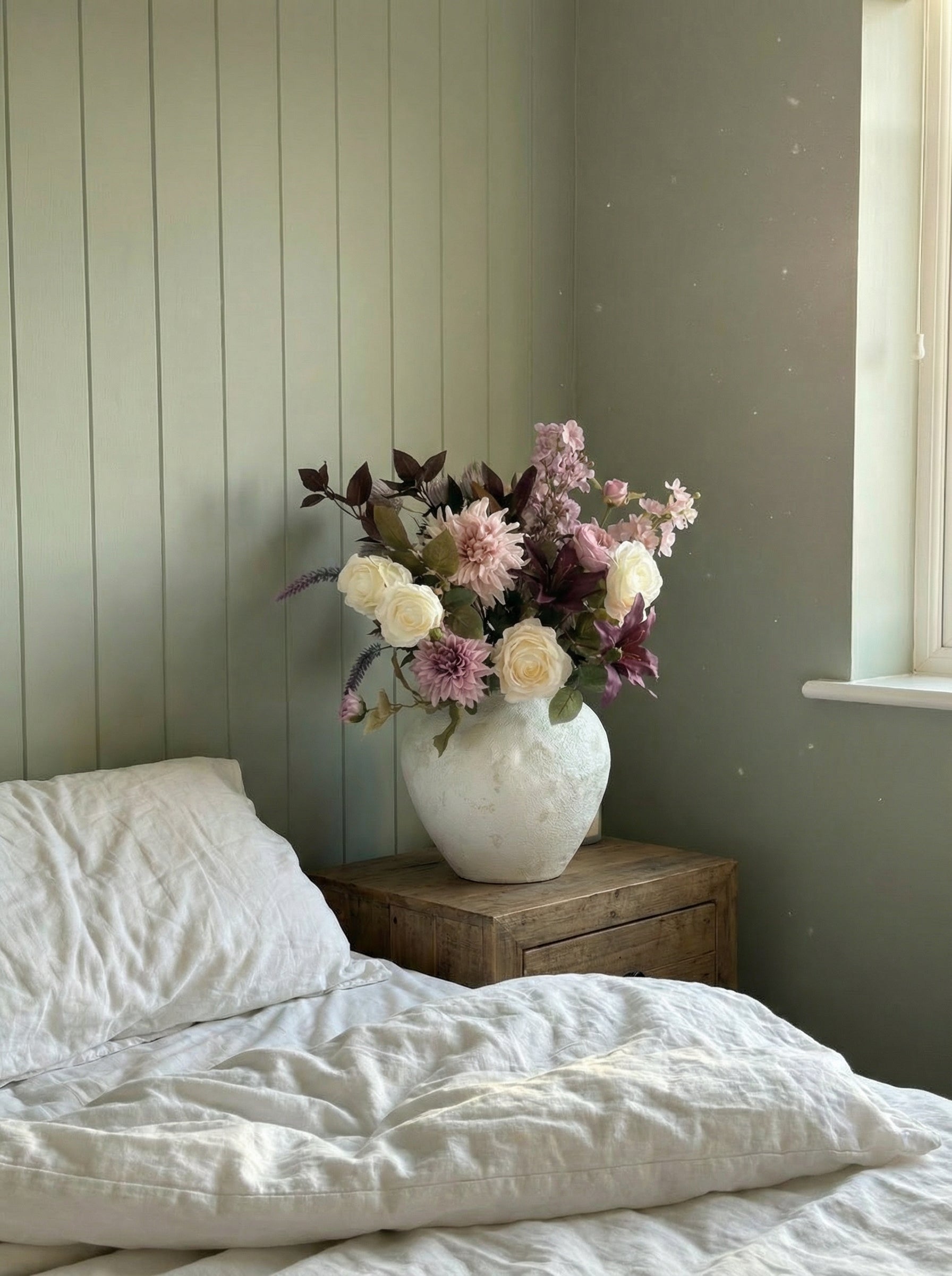 Floral arrangement in a white vase on a wooden nightstand next to a bed with white bedding.