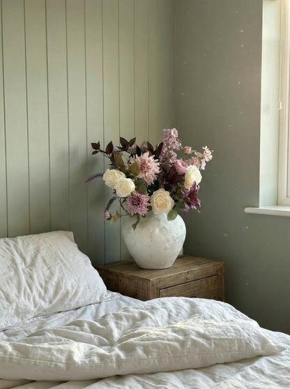 Floral arrangement in a white vase on a wooden nightstand next to a bed with white bedding.
