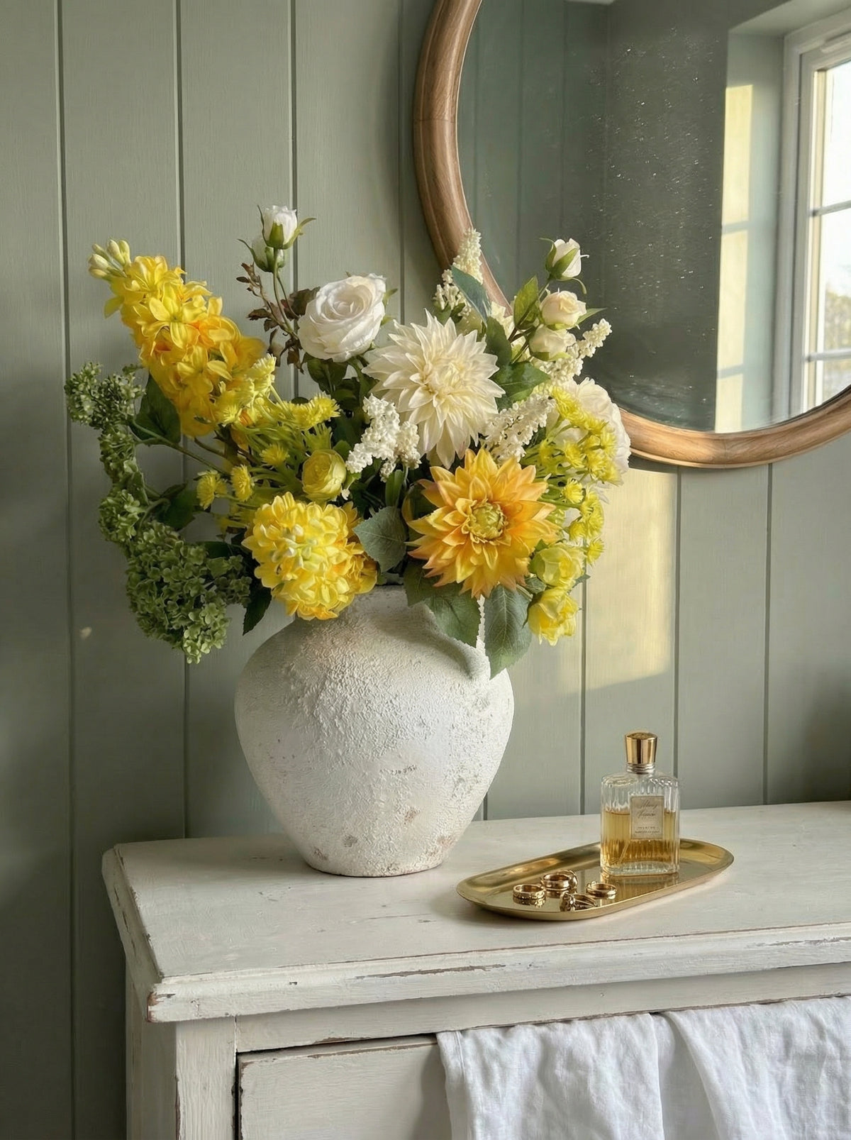 Vase of yellow and white flowers on a wooden surface with a mirror and perfume bottle in the background.