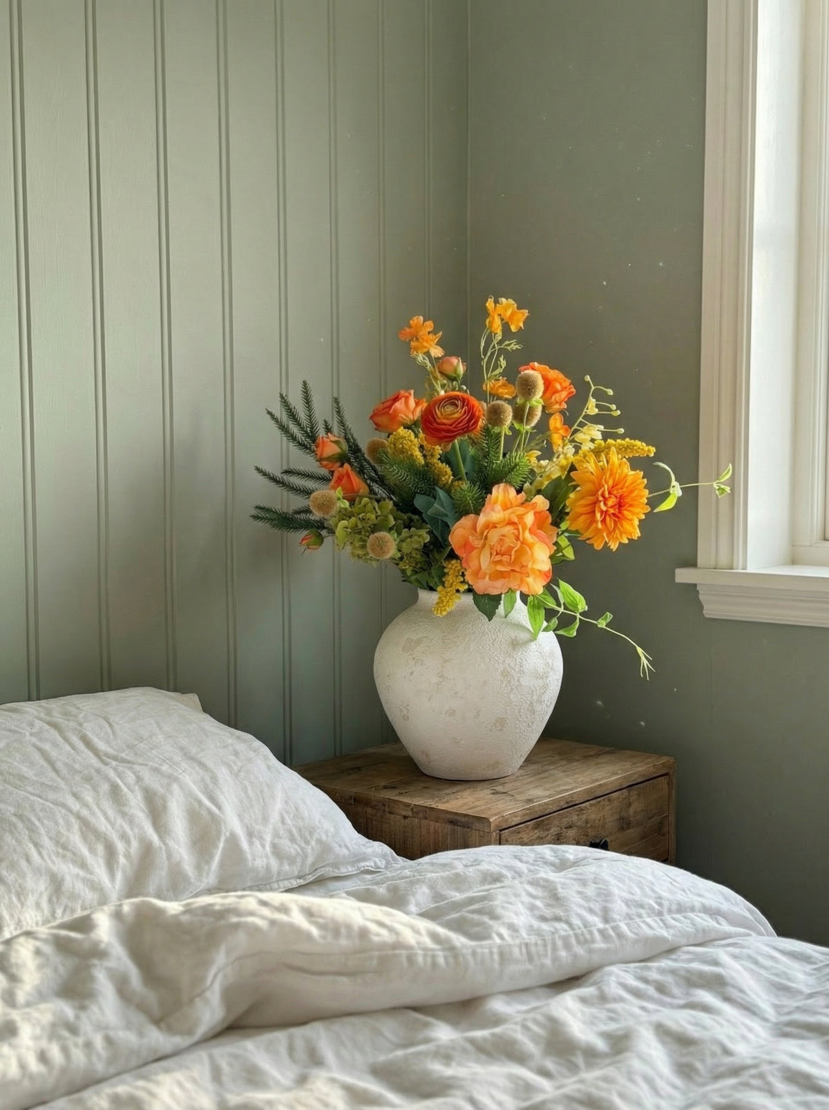 Vase with flowers on a wooden nightstand next to a bed in a room with green walls.