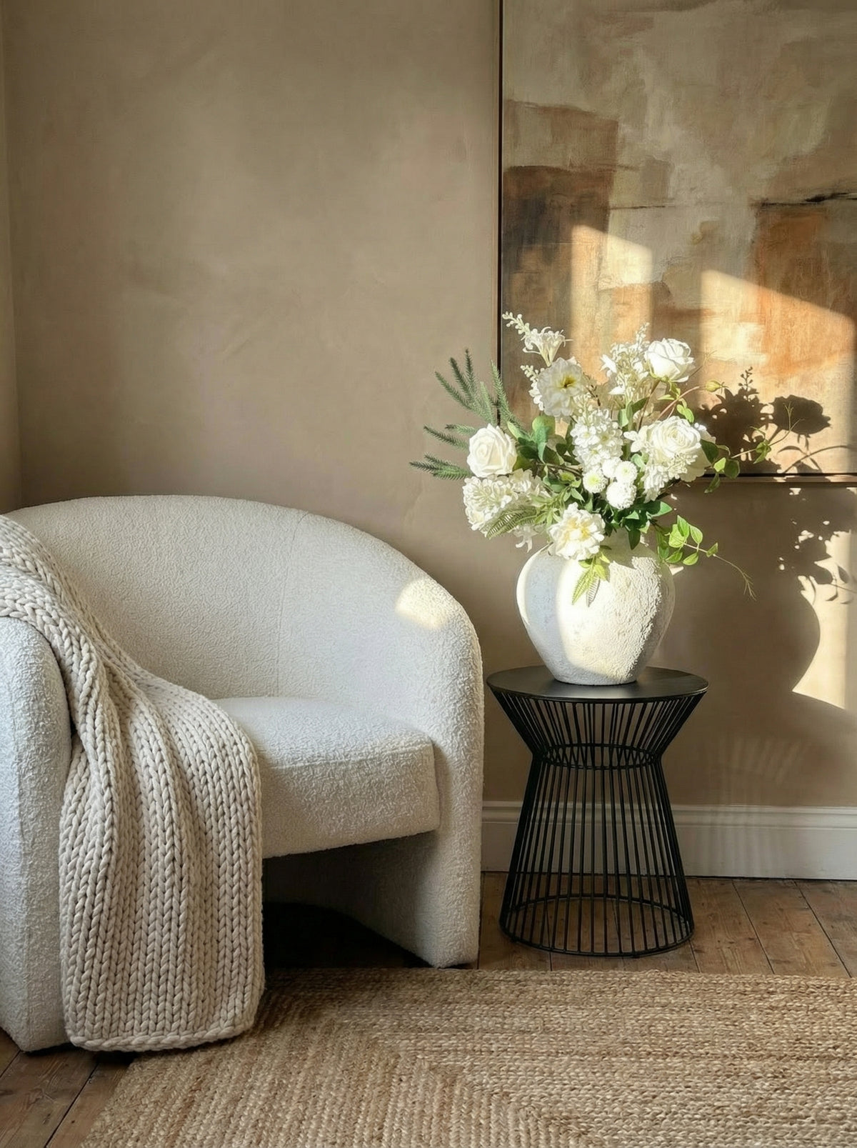 White armchair with a textured blanket and a vase of white flowers on a black wire table against a beige wall.
