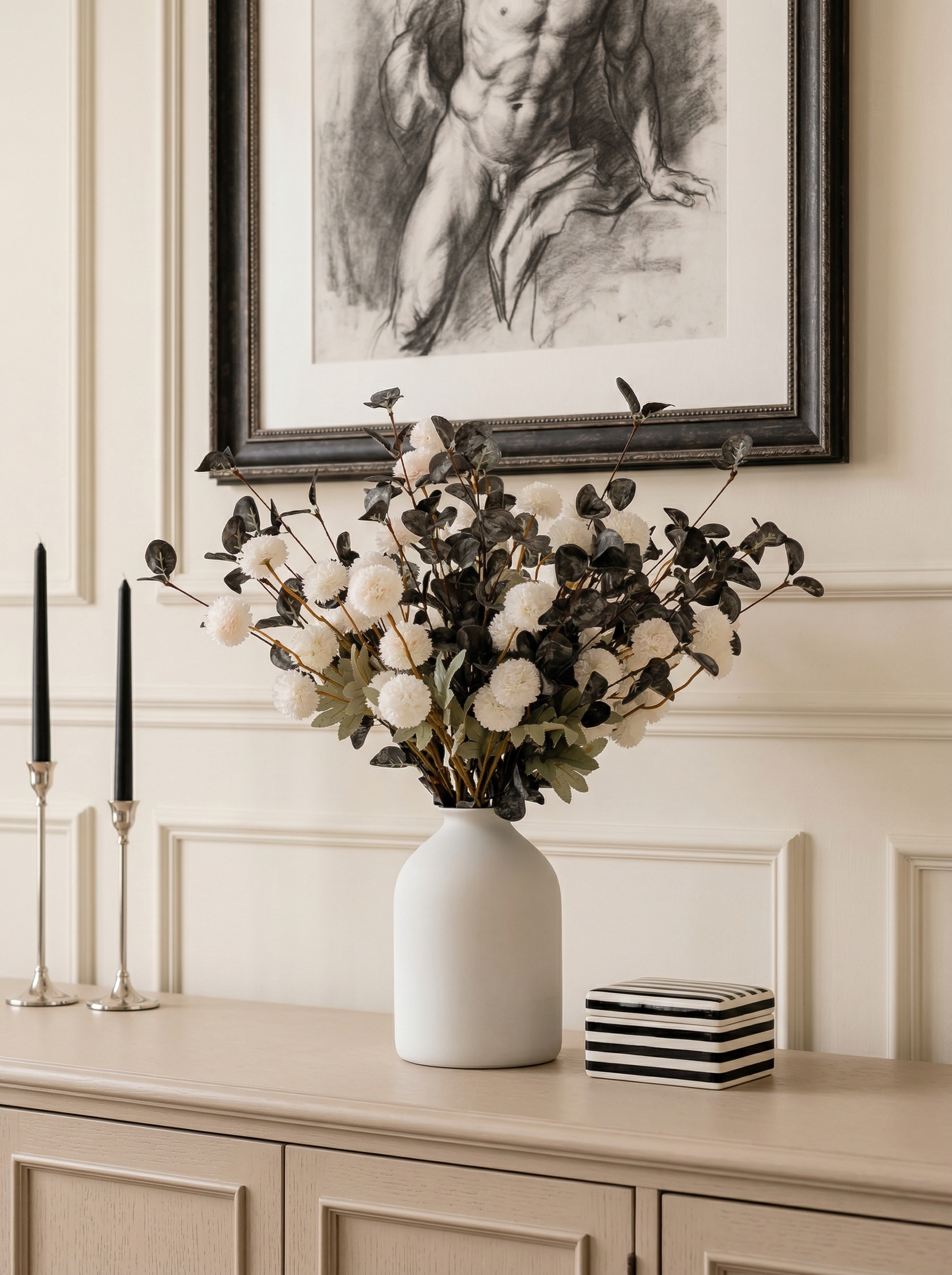 White vase with flowers on a wooden cabinet against a wall with a framed black and white artwork.