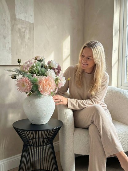 Woman sitting on a chair next to a vase of flowers in a room with light-colored walls.