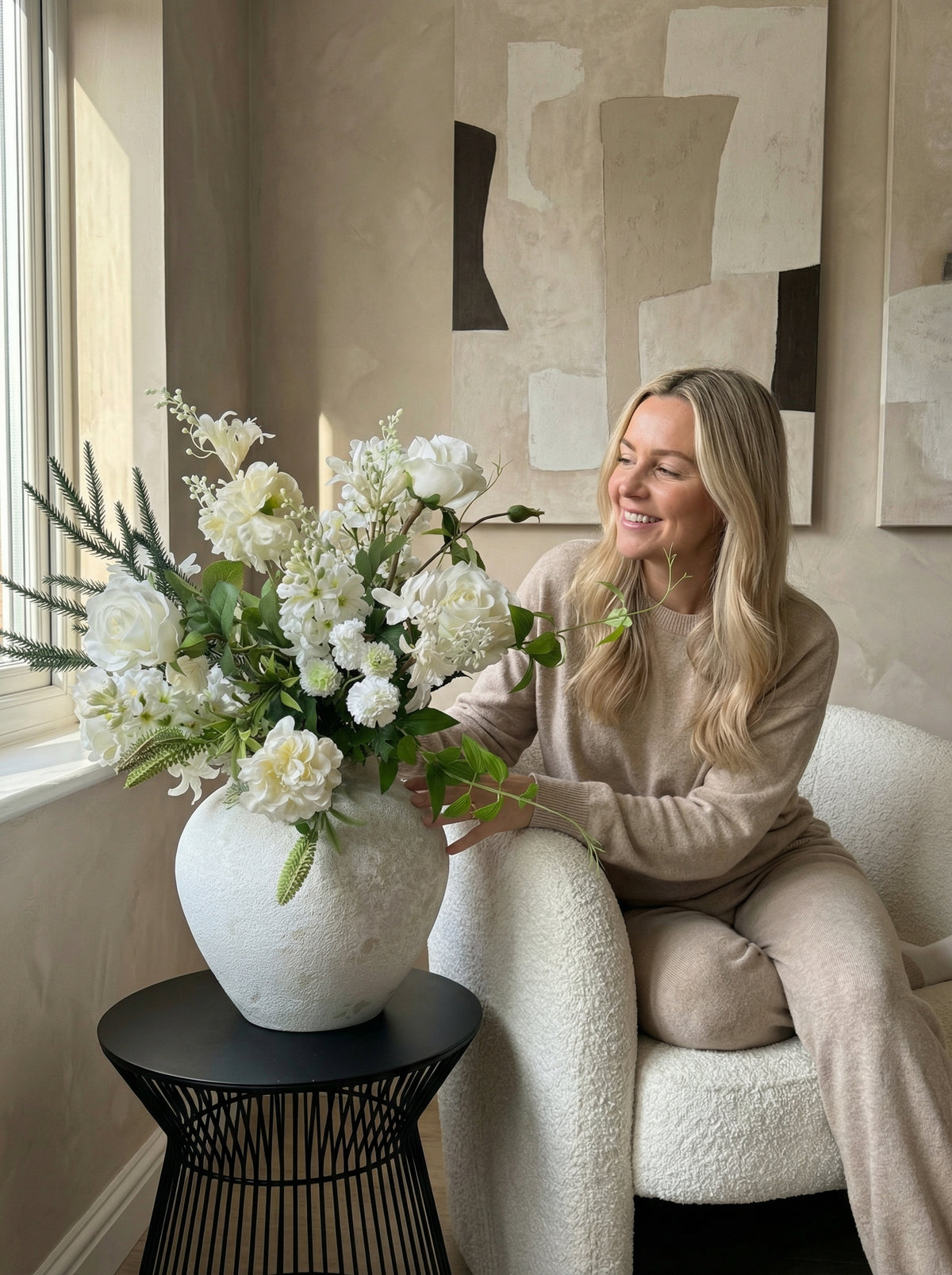 Woman sitting on a couch with a large vase of white flowers in a stylish room.