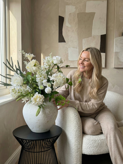 Woman sitting on a couch with a large vase of white flowers in a stylish room.