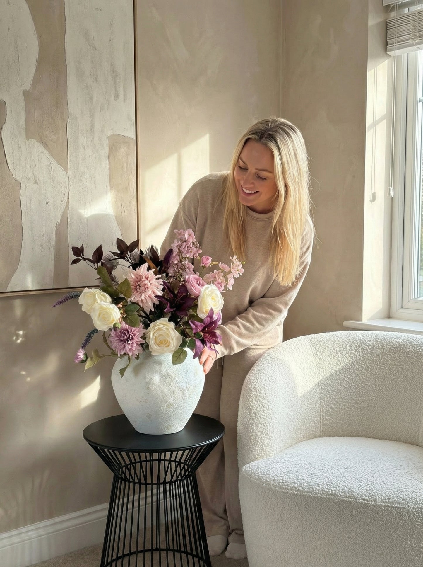 Woman sitting on a white chair next to a vase of flowers in a room with neutral decor.