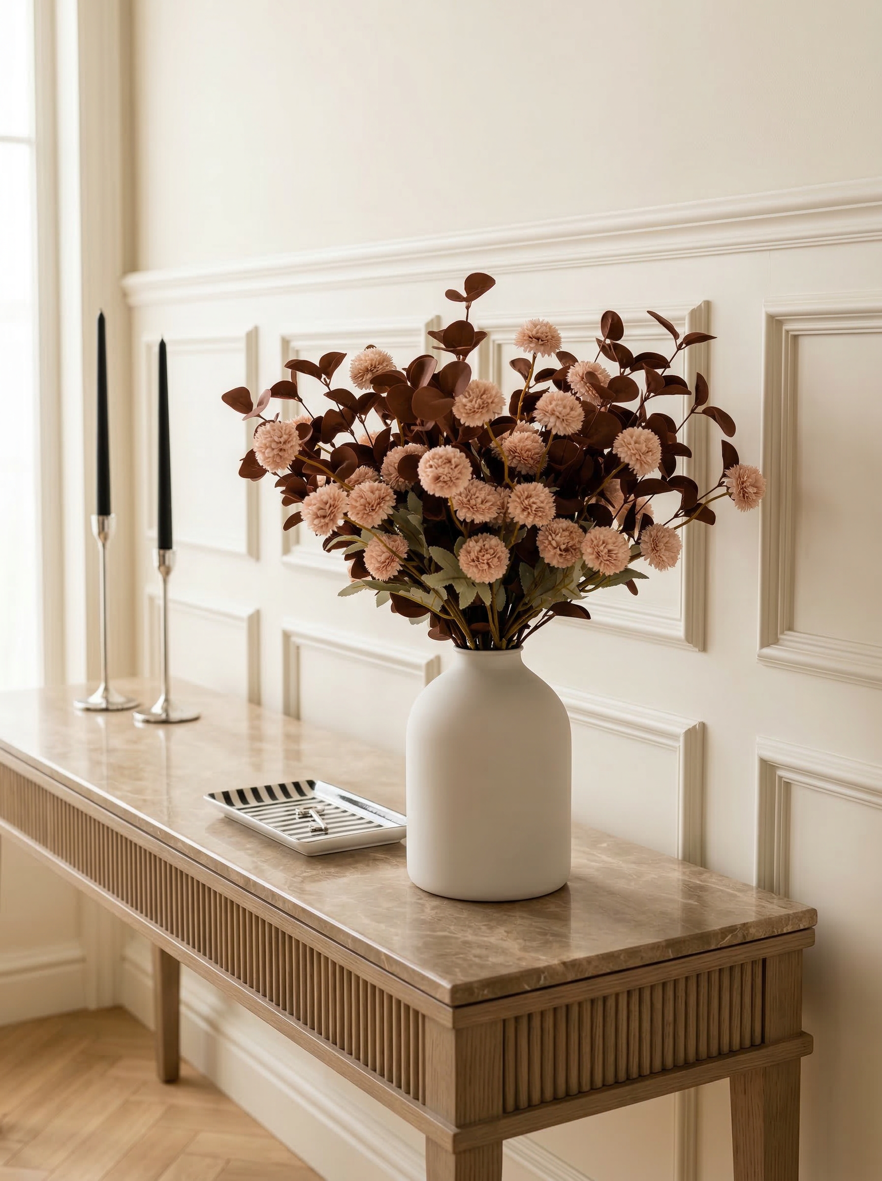 White vase with pink flowers on a wooden console table against a white paneled wall.